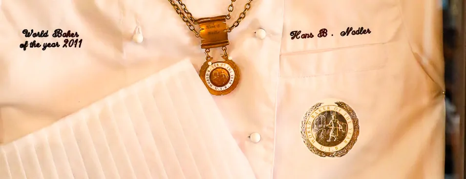 Close-up of the framed chef’s jacket: “World Baker of the year 2011” embroidered on one side, “Hans B. Nadler” on the other, a medal between them.