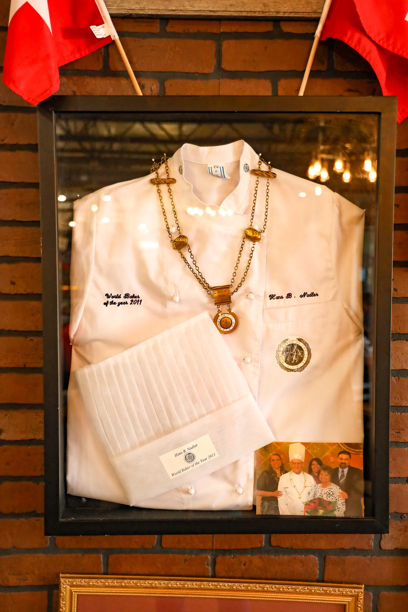 Hans B. Nadler’s white chef’s jacket and toque framed on a brick wall, with a medal on a chain, embroidery reading “World Baker of the year 2011” and a family photograph.