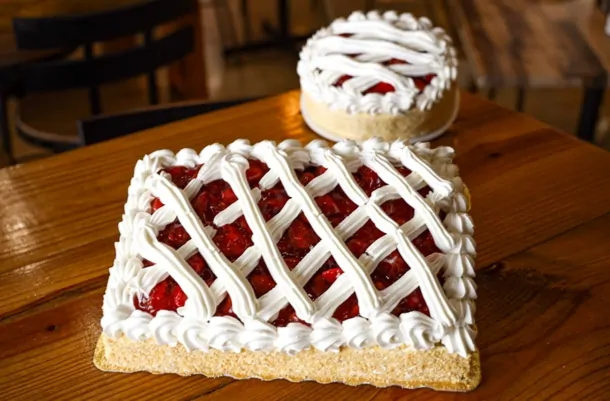 A rectangular cake topped with cherries under a lattice of piped cream, a round one behind it.