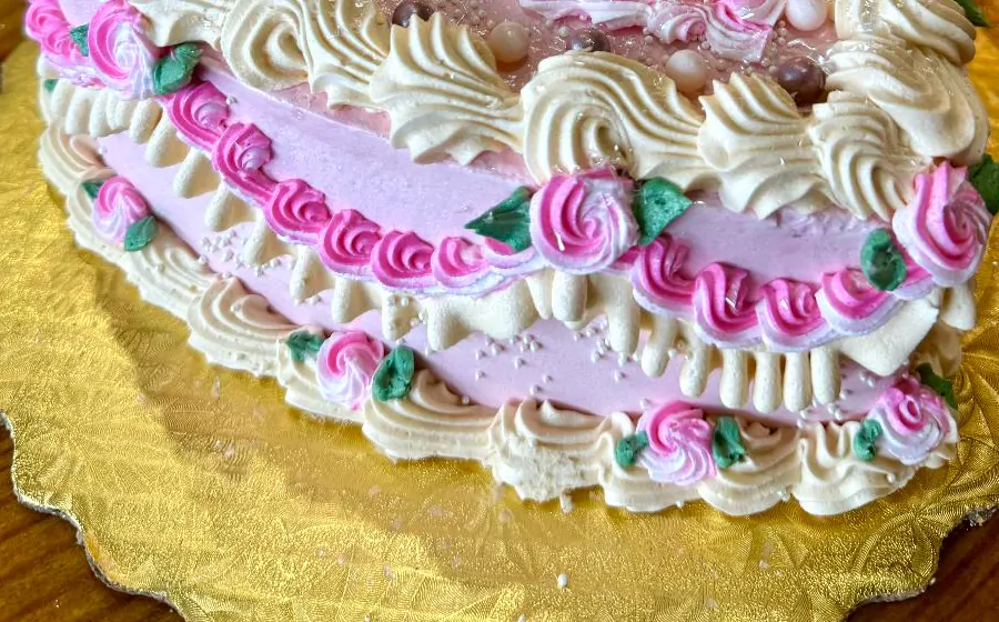 Close-up of the side of a heart cake: pink rosettes with green leaves between rows of piped cream.