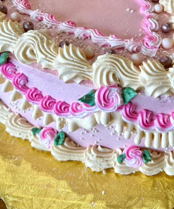 Close-up of a pink heart cake: cream shells and sugar pearls along the top, pink rosettes with green leaves around the side, on a gold scalloped board.