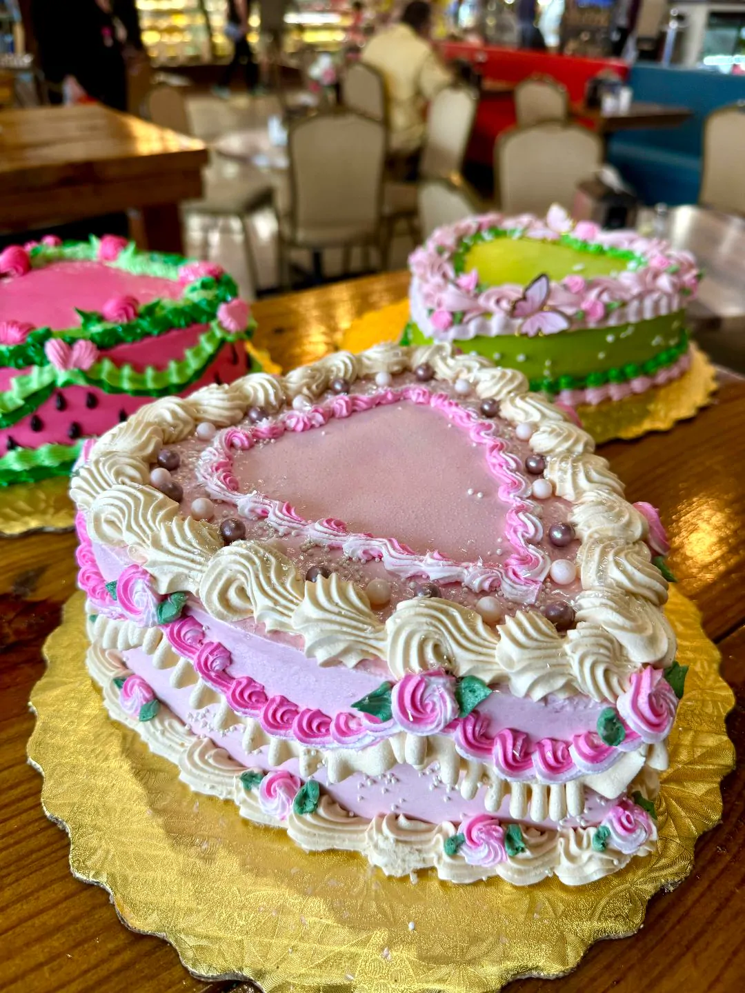 Three heart-shaped cakes on wooden tables: pink with cream piping in front, a watermelon design and a green cake with a butterfly behind.
