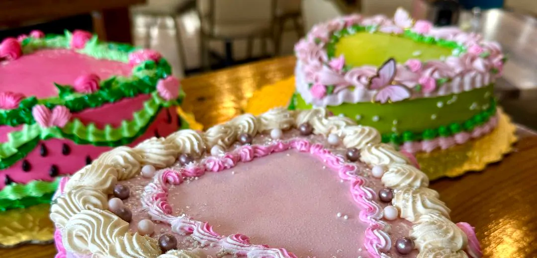 A watermelon-patterned heart cake and a green heart cake with a pink butterfly, softly out of focus.