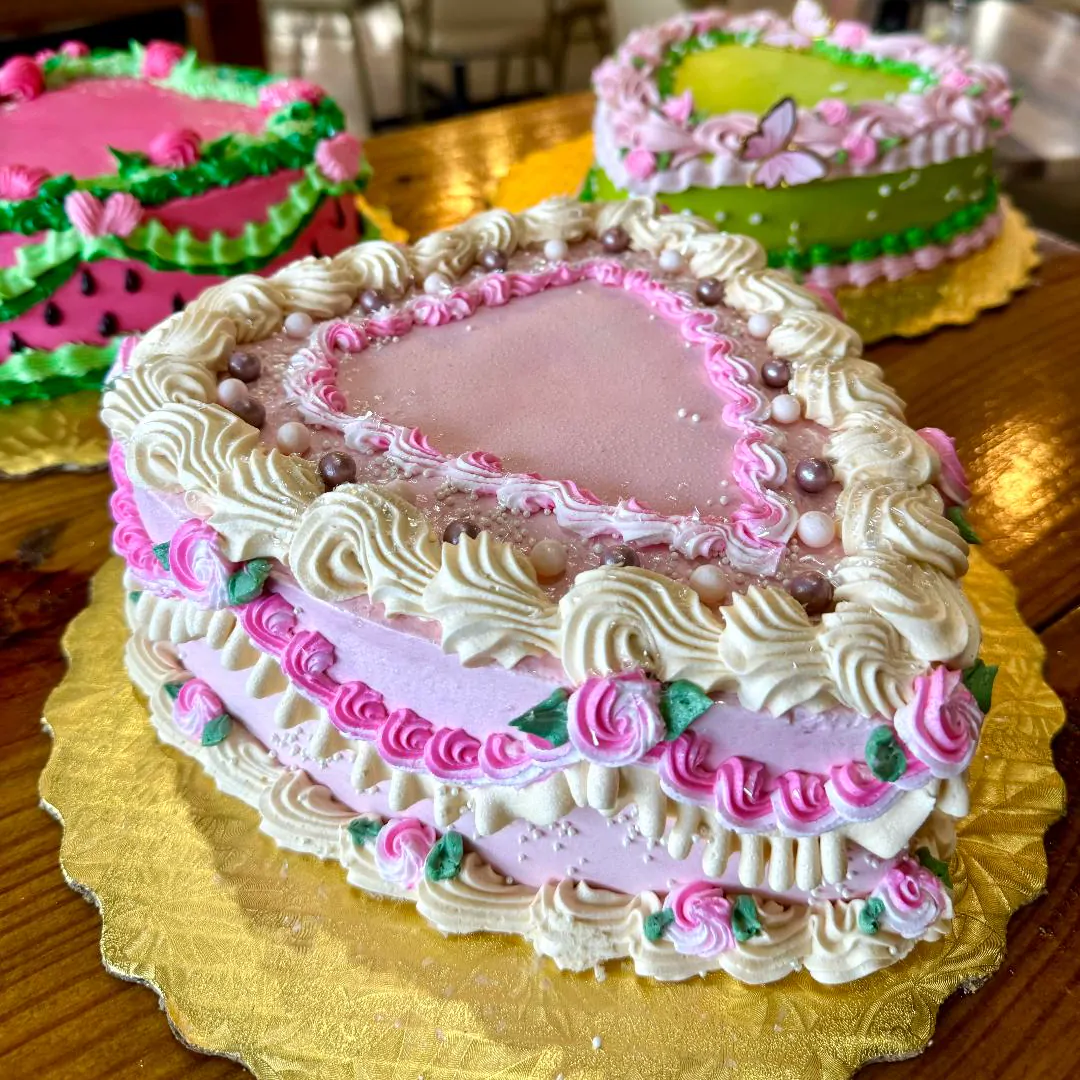 A pink heart-shaped cake with piped cream shells and rosettes on a gold scalloped cake board, two more heart cakes behind it.