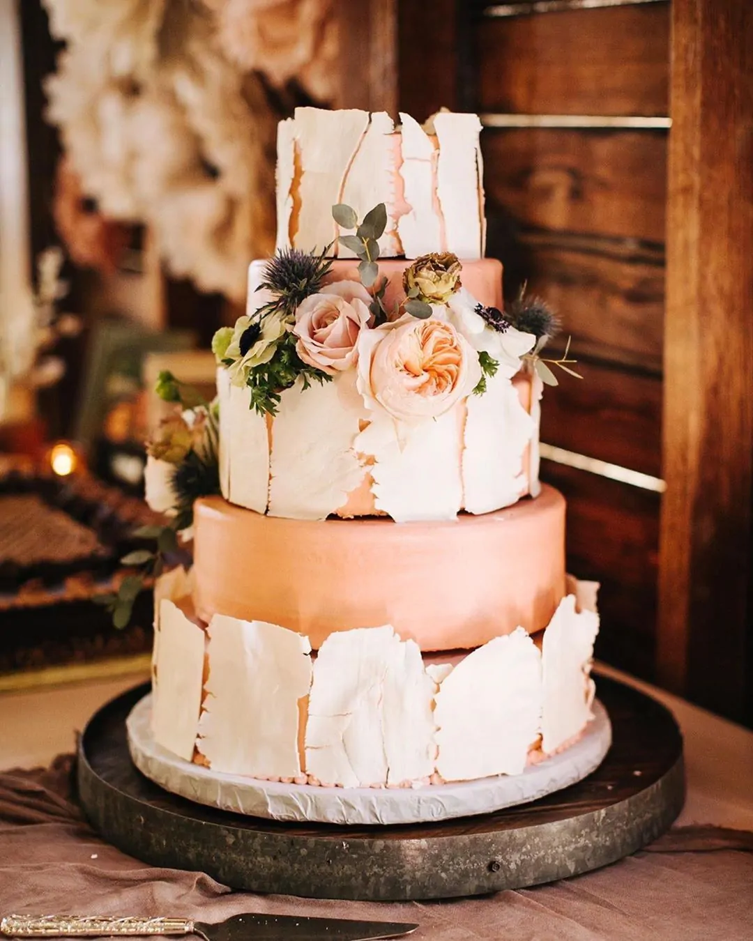 A tiered wedding cake alternating peach tiers with tiers wrapped in white shards, finished with roses, thistle and greenery.